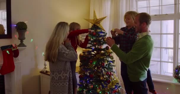 幼い息子と娘を抱えている白人のカップルの側の景色とクリスマスの時に彼らのシッティングルームで一緒にクリスマスツリーを飾る — ストック動画