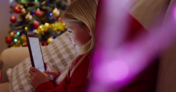 クリスマスの時間にソファの上に座っているタブレットを使用して若い白人の少女の側面図 背景に装飾されたクリスマスツリーと前景に輝く妖精の光 — ストック動画