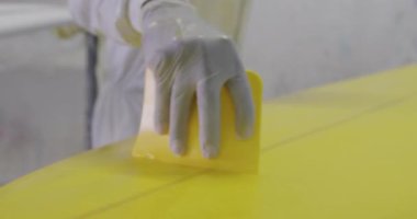 Atölyesindeki bin yıllık beyaz erkek sörf tahtasının orta kesiminde koruyucu giysiler ve eldivenler giyip, sörf tahtasında bir araç kullanarak üretim sürecinde bir kaplama uyguluyor.