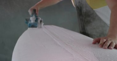 Atölyesinde bin yıllık beyaz bir erkek sörf tahtası üreticisinin yan görüntüsü. Üretim sırasında yeni bir sörf tahtasını bir aletle zımparalıyor.