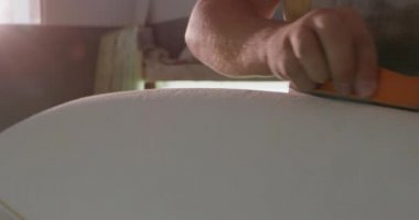 Atölyesinde bin yıllık beyaz bir erkek sörf tahtasının ön tarafı. Üretim sırasında yeni bir sörf tahtasının kenarlarını bir aletle ovuyor.