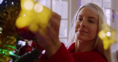 Gülümseyen sarışın beyaz bir kadının evinin oturma odasındaki Noel ağacını süslediği ön manzara.