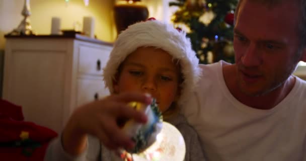 크리스마스 시간에 자신의 거실에서 스노우 글로브를 모자를 아들과 바닥에 앉아있는 — 비디오