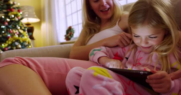 一位快乐的白人年轻母亲坐在沙发上 带着年幼的女儿在客厅里用平板电脑 圣诞节前夕 母亲在客厅里爱抚着女儿 — 图库视频影像
