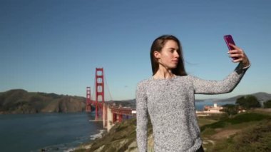Önde, arkasında Golden Gate Köprüsü ve San Francisco Körfezi Bölgesi olan akıllı telefon kullanarak uzun siyah saçlı ve selfie çeken beyaz bir kadın var.