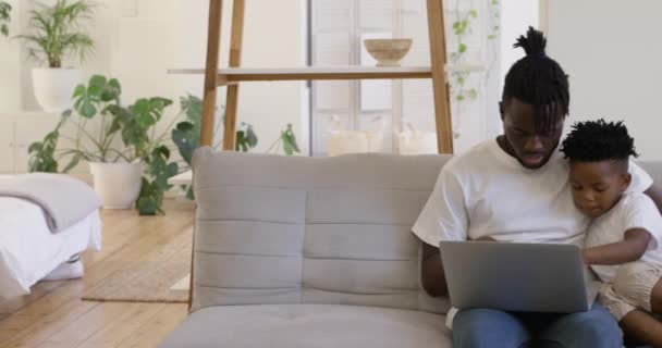 一个微笑的千年非洲裔美国父亲和他年幼的儿子在家里 坐在客厅的沙发上 一起用笔记本电脑聊天 — 图库视频影像