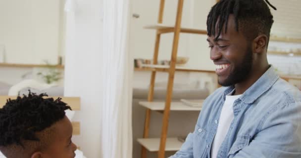 彼は家でベッドに座っている間 彼の若い息子が服を着るのを助ける笑顔の千年のアフリカ系アメリカ人の父親の近くにサイドビュー スローモーション — ストック動画