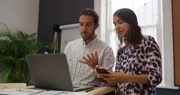 在一个创意企业的办公室里工作的一千年白人男女坐在办公桌前 用笔记本电脑 女人给男人看她的智能手机 — 图库视频影像