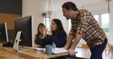 Bin yıllık beyaz bir adam ve iki kadının yaratıcı bir iş ofisinde çalışırken, konuşurken ve masaüstü bilgisayarlarını kullanırken yan görüntüsü