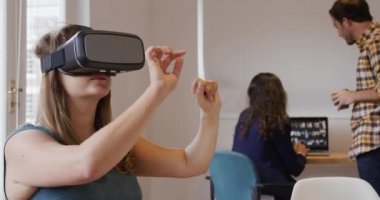 Bin yıllık beyaz bir kadının yaratıcı bir iş ofisinde çalıştığını, bir masada VR kulaklıkla oturduğunu ve arka planda çalışan iki iş arkadaşıyla ellerini önünde gezdirdiğini.