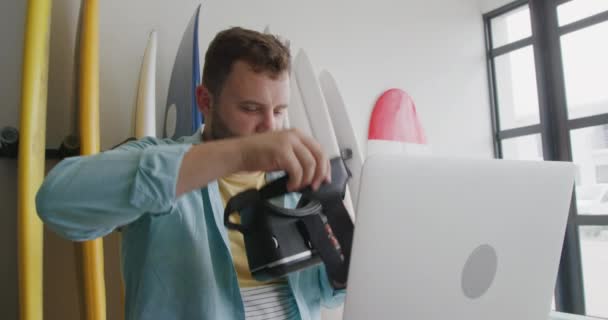 ノートパソコンを使って机に座ってVrヘッドセットに彼の後ろにサーフボードのラックを置く彼のスタジオで百万人の白人男性サーフボードメーカーの近くのフロントビュー — ストック動画