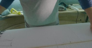 Sörf tahtasını inceleyen, kameraya gülümseyen ve kollarını kavuşturan bin yıllık beyaz bir erkek sörf tahtası yapımcısının portresi.