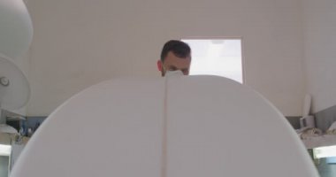 Atölyesinde yüz maskesi takan ve üretim sırasında sörf tahtasını yakından inceleyen bin yıllık beyaz bir erkek sörf tahtasının ön görüntüsü.