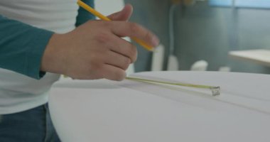 Atölyesinde bin yıllık beyaz erkek sörf tahtasının orta kesiminde üretim sırasında yeni bir sörf tahtasını ölçüyor ve işaretliyor.