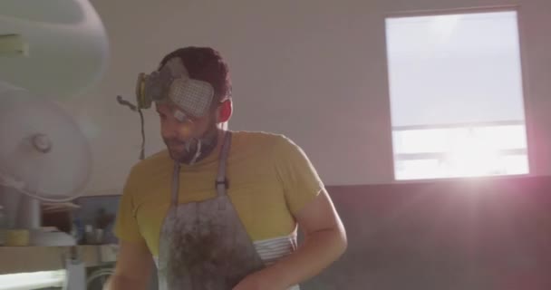 Vue de face d'un fabricant de planches de surf blanc millénaire dans son studio, ponçant une planche de surf avec un outil, un masque respirateur sur le dessus de sa tête 