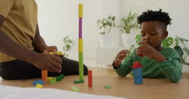Bin yıllık Afro-Amerikan bir baba ve genç oğlunun yan görüntüsü. İnşaat taşlarıyla oynuyorlar. Onları evdeki bir masada dengede tutuyorlar.