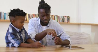 Bin yıllık Afro-Amerikan bir baba ve genç oğlunun ön planda yan yana oturması, babanın elinde küçük bir dron tutması ve ikisinin de masaya bakması, tablet bir bilgisayar, yavaş çekim.