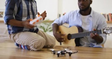 Bin yıllık Afro-Amerikan bir baba ve genç oğlunun birlikte evde akustik gitar çalarken, genç oğlu elinde bir cep telefonu ve ağır çekimde küçük bir drone ile masada otururken ön plan görüntüsü.