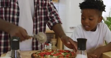Binlerce yıllık gülümseyen Afrikalı bir Amerikalı baba ve oğlunun evdeki mutfakta birlikte yemek hazırlayıp konuşmalarının ön planında, baba, oğlu izlerken pişirdikleri pizzayı kesiyor.