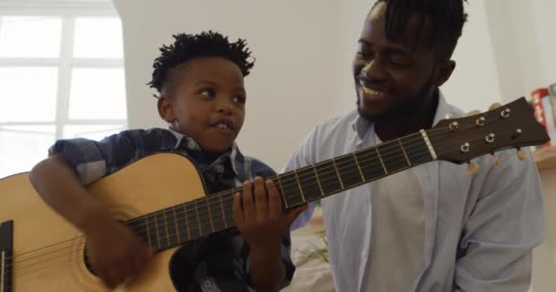 彼の千年の父は彼の笑顔の後ろに座っている間 フロントビューは自宅でアコースティックギターを演奏若いアフリカ系アメリカ人の少年のクローズアップ スローモーション — ストック動画