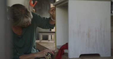 Ön planda gülümseyen beyaz bir kadın var. Evdeki bir atölyede tornavida kullanıyor ve ağır çekimde mobilya monte ediyor.