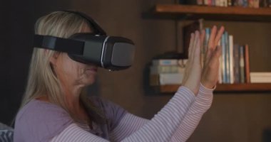 Oturma odasında VR kulaklık takan yetişkin beyaz bir kadının yan görüntüsü. Ellerini kaldırıyor, el sallıyor, sallıyor ve etrafa bakıyor. Yavaş çekim.