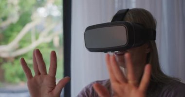 Oturma odasında VR kulaklık takan mutlu, beyaz bir kadının ön görüntüsü. Ellerini kaldırıyor ve gülümsüyor, ağır çekimde etrafa bakıyor.