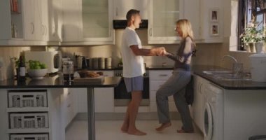 Mutfakta çıplak ayakla dans eden ve sabah güneşinde gülümseyen beyaz bir çiftin yan görüntüsü, yavaş çekim.