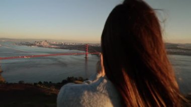 Golden Gate Ulusal Rekreasyon Bölgesi 'nde dikilen beyaz saçlı genç bir kadının arka görüntüsü. Golden Gate Köprüsü ve San Francisco Körfezi bölgesinin resimlerini çekiyor. Akıllı telefonuyla, ağır çekimde.