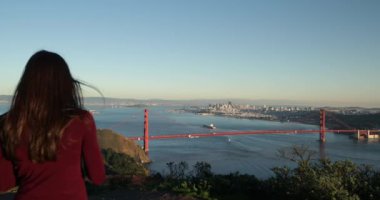 Golden Gate Ulusal Dinlenme Alanı 'nda yürüyen ve Golden Gate Köprüsü' nün ve San Francisco Körfezi Bölgesi 'nin akıllı telefonuyla fotoğraflarını çeken beyaz saçlı genç bir kadının arka görüntüsü.