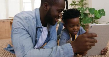 Binlerce yıllık gülümseyen Afro-Amerikan bir adam ve genç oğlunun oturma odalarında birlikte tablet bilgisayar kullanarak yere yatışının ön görüntüsü.