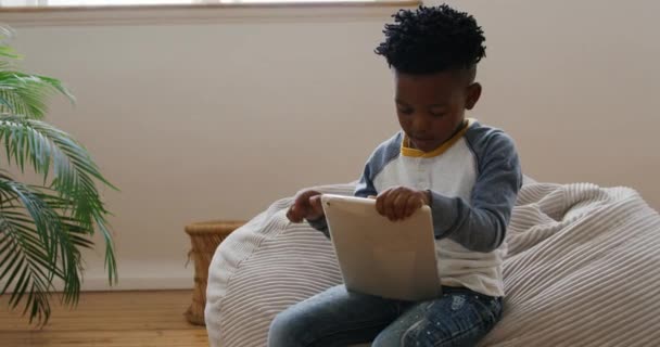 タブレットコンピュータを使って静かに豆袋の上に座っている若いアフリカ系アメリカ人の少年のフロントビュー スローモーション — ストック動画