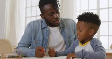Bin yıllık Afro-Amerikan bir babanın ve küçük oğlunun birlikte evde bir masada oturup pastel boyalarla oynayıp, konuşup, ödev yapmasının ön görüntüsü.
