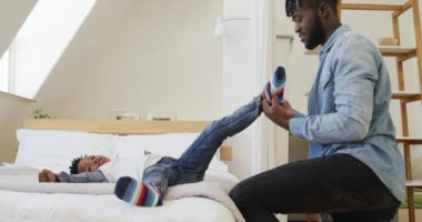 Binlerce yıllık gülümseyen Afro-Amerikan bir babanın, genç oğluna çoraplarını giydirmesine yardım ederken evinde ağır çekimde yatışının yan görüntüsü.