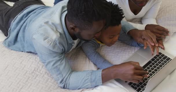 一个千禧年的非洲裔美国家庭和他们年幼的儿子在家里一起躺在床上 他们用一台笔记本电脑 父亲用笔记本电脑帮助儿子打字 动作缓慢 — 图库视频影像