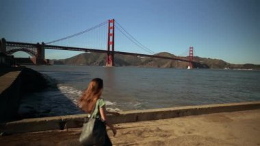 San Francisco Körfez Bölgesi 'ndeki Golden Gate Köprüsü' nün yanında uzun siyah saçlı genç beyaz bir kadının arka görüntüsü ve akıllı telefonunu kullanmak için durması, ağır çekim.