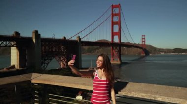 Arka planda, uzun siyah saçlı, akıllı telefonuyla selfie çeken beyaz bir kadın, Golden Gate Köprüsü ve San Francisco Körfezi Bölgesi, yavaş çekim.