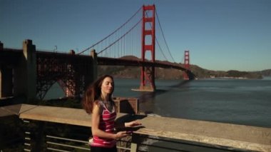 Arka planda Golden Gate Köprüsü ve San Francisco Körfez Bölgesi ile özçekim yapmak için akıllı telefonunu kullanan uzun siyah saçlı beyaz bir kadının ön görüntüsü.