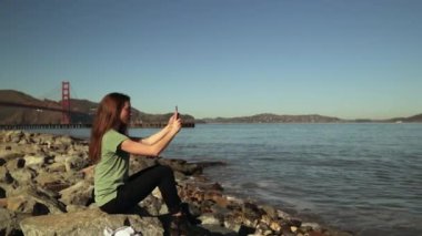 Suyun kenarındaki kayalarda oturan, Golden Gate Köprüsü ve arka planda San Francisco Körfezi Bölgesi olan akıllı telefon kullanarak selfie çeken uzun siyah saçlı bir kadının yan görüntüsü.