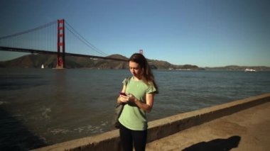 Arka planda Altın Kapı Köprüsü ve San Francisco Körfezi Bölgesi olan akıllı telefon kullanarak uzun siyah saçlı beyaz bir kadının ön görüntüsü, yavaş çekim.