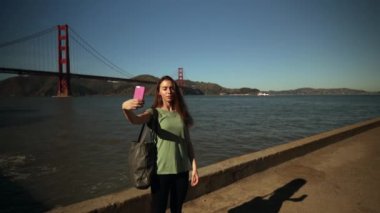 Arka planda Golden Gate Köprüsü ve San Francisco Körfez Bölgesi olan akıllı telefon kullanarak uzun siyah saçlı ve selfie çeken bir kadının ön görüntüsü, yavaş çekim.