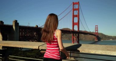 Arka planda, Golden Gate Ulusal Eğlence Bölgesi, San Francisco Körfezi Bölgesi ile Golden Gate Köprüsü 'nün fotoğrafını çekmek için akıllı telefonunu kullanan uzun siyah saçlı beyaz bir kadın var.