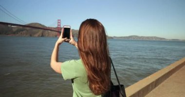 Arkadaki görüntüde, uzun siyah saçlı, Golden Gate Köprüsü ve San Francisco Körfezi bölgesinin fotoğraflarını çeken beyaz bir kadın telefonuna bakıyor ve gülümsüyor.
