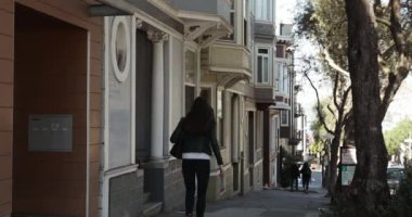 Arkadaki görüntüde, elinde çanta taşıyan bir kadın ağaçlarla dolu bir şehir sokağında yürüyor. Kameradan uzaklaşıyor..