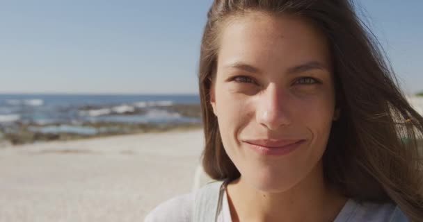 明るいビーチでカメラに笑みを浮かべて長い暗い髪を持つ幸せな若い白人女性の肖像画のクローズアップ スローモーション — ストック動画