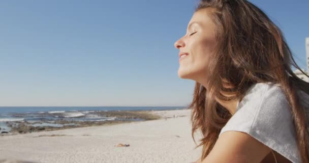 一个快乐的白人年轻女子靠着篱笆坐在阳光灿烂的海滩上 微笑着转向镜头 慢吞吞地朝旁边看去 — 图库视频影像