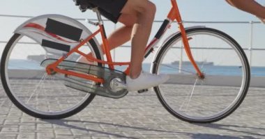 Güneşli bir günde deniz kenarında bisiklet sürerken eğlenen genç, beyaz bir çiftin yan görüntüsü. Kadın bacaklarını kaldırmış, ağır çekimde gidona otururken pedal çeviren adam.