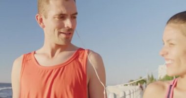 Önde, spor giyimli, mutlu, genç, beyaz bir çiftin, güneşin altında yürüyüp konuştuğu ve bir çift kulaklığı paylaştığı görüntüler.