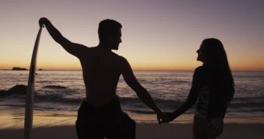 Deniz kenarında öpüşen, gün batımına karşı siluetlenmiş, sörf tahtasına yaslanmış, ağır çekim, mutlu, genç bir beyaz çiftin yan görüntüsü.