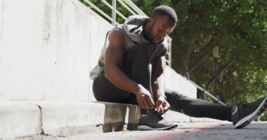 Afrika kökenli Amerikalı bir adamın kolsuz kapüşonlu ve spor giysili bir adamın güneşli bir günde şehirde antrenman yapmadan önce bir basamağa oturup ayakkabı bağcıklarını bağlarken görüntüsü, yavaş çekim.